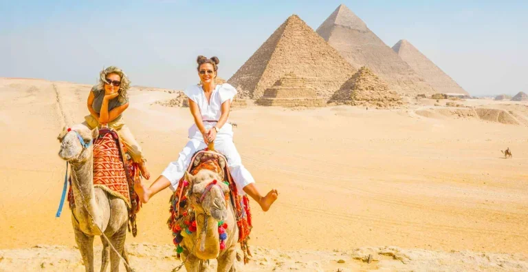 Two smiling female tourists riding camels in the Giza desert, with the Great Pyramids in the background on an Egypt Day Tours excursion.